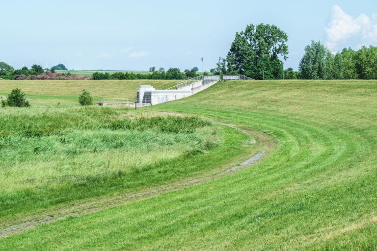 Das Bild zeigt eine grüne Landschaft mit einer Hochwasserschutzanlage. Die Anlage besteht aus einer Betonmauer und einem Tor, das auf einer erhöhten Fläche liegt. Ein geschwungener Weg führt zum Staudamm, umgeben von Wiesen und Bäumen im Hintergrund.