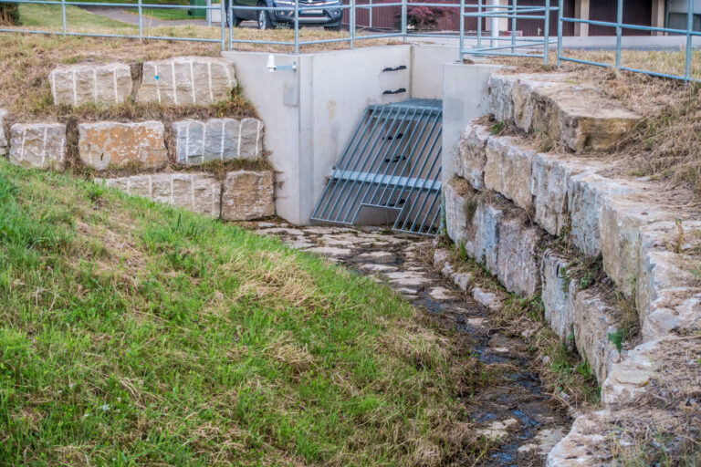 Das Bild zeigt eine Hochwasserschutzanlage mit einem Betonkanal, an dessen Eingang ein Metallgitter angebracht ist, um Treibgut fernzuhalten. Die Umgebung ist mit großen Steinblöcken gesichert, um Erosion zu verhindern und die Ufer zu stabilisieren. Auf der Anlage befindet sich ein Metallgeländer als Sicherheitsmaßnahme.