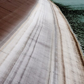 Das Bild zeigt eine geneigte Betonfläche mit deutlich sichtbaren horizontalen Linien, die auf unterschiedliche Wasserstände im Laufe der Zeit hinweisen. Rechts im Bild befindet sich ein Gewässer mit kleinen Wellen auf der Oberfläche. Die linke Seite der Betonfläche ist trocken und weist eine rötlich-braune Färbung auf.