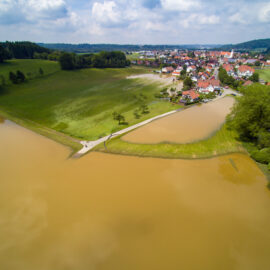 Das Bild zeigt eine Luftaufnahme einer Kleinstadt mit einem braun gefärbten Gewässer im Vordergrund, einer Überschwemmung. Umgeben ist die Stadt von grünen Feldern, Baumgruppen und sanften Hügeln im Hintergrund die teilweise überflutet wurden.