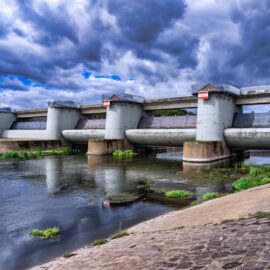 Das Bild zeigt eine Staumauer mit mehreren zylindrischen Pfeilern, die eine brückenartige Plattform tragen. Der Damm befindet sich über einem Kanal, im Vordergrund führt eine gepflasterte Fläche zum Wasser hinunter. Der Himmel ist von dunklen, dramatischen Wolken bedeckt, was auf ein bevorstehendes Unwetter hindeutet.