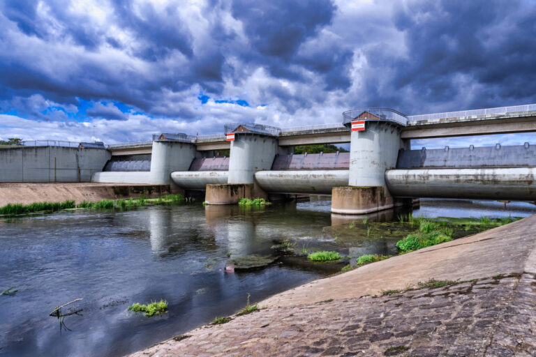 Das Bild zeigt eine Staumauer mit mehreren zylindrischen Pfeilern, die eine brückenartige Plattform tragen. Der Damm befindet sich über einem Kanal, im Vordergrund führt eine gepflasterte Fläche zum Wasser hinunter. Der Himmel ist von dunklen, dramatischen Wolken bedeckt, was auf ein bevorstehendes Unwetter hindeutet.