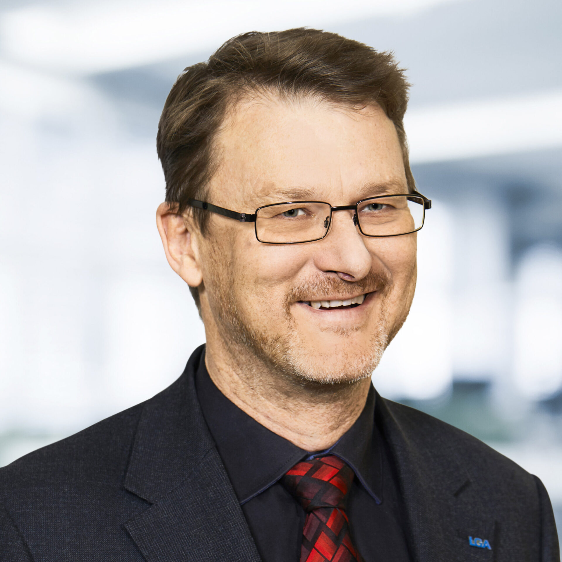 Manfred Steinicke trägt eine Brille, einen dunklen Anzug, ein dunkles Hemd und eine rot-schwarz karierte Krawatte. Der Hintergrund ist unscharf und zeigt ein Büro.