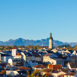 Malerischer Blick auf Traunstein mit roten Ziegeldächern, einem markanten Kirchturm im Zentrum und den Alpen im Hintergrund unter klarem, blauem Himmel an einem sonnigen Tag.