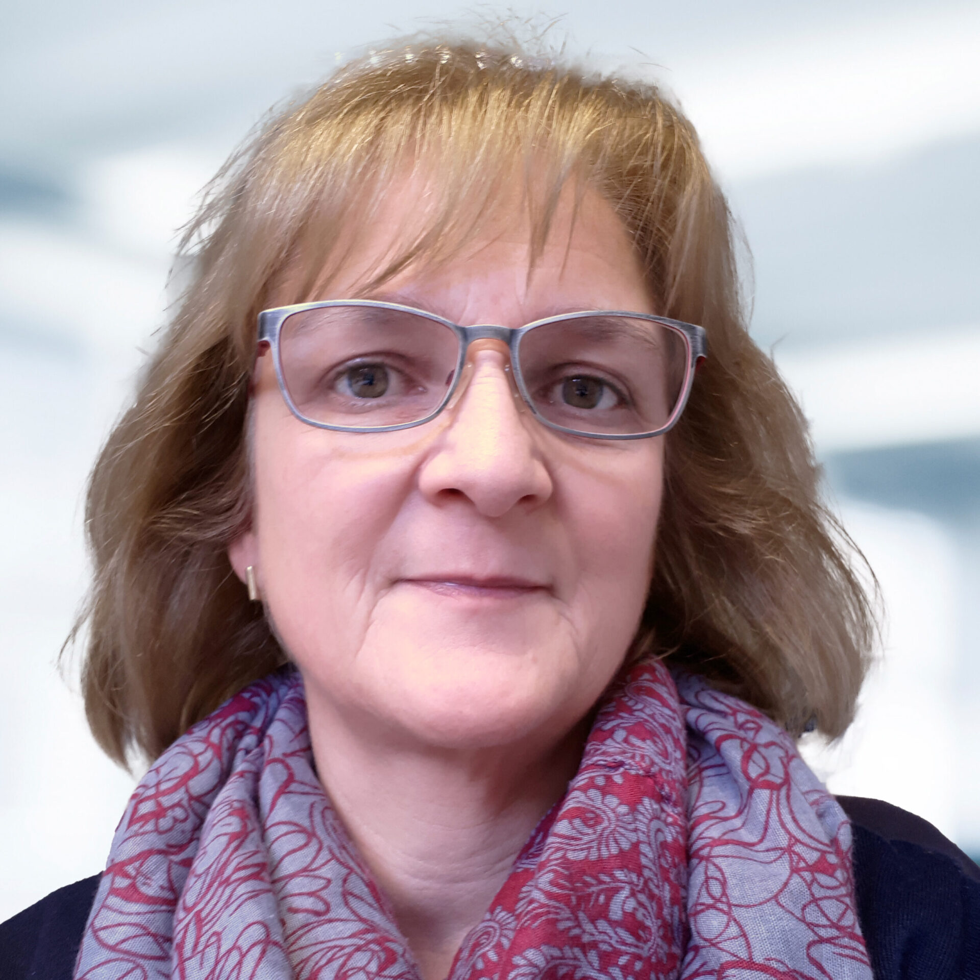 Heike Trautner trägt eine Brille und ein Tuch mit rotem Muster. Der Hintergrund ist unscharf und zeigt ein Büro.