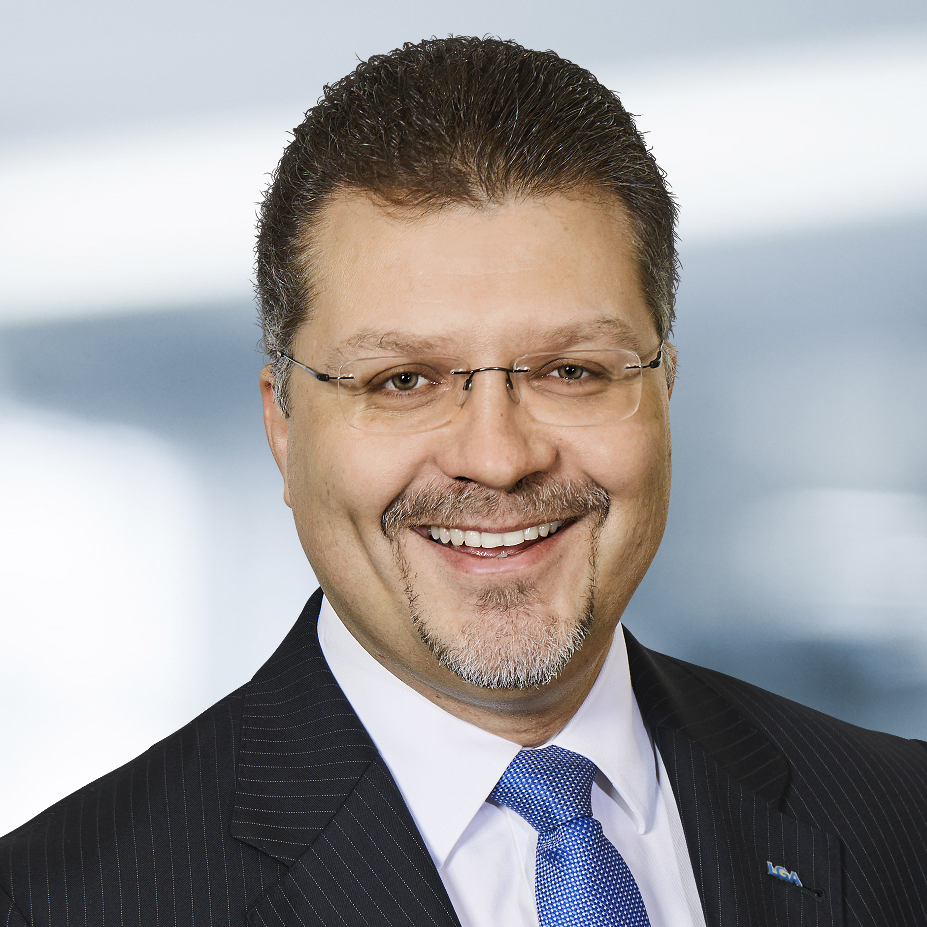 Hans-Peter Trinkl trägt einen dunklen Nadelstreifenanzug, ein weißes Hemd und eine blaue Krawatte. Der Hintergrund ist unscharf und zeigt ein Büro.