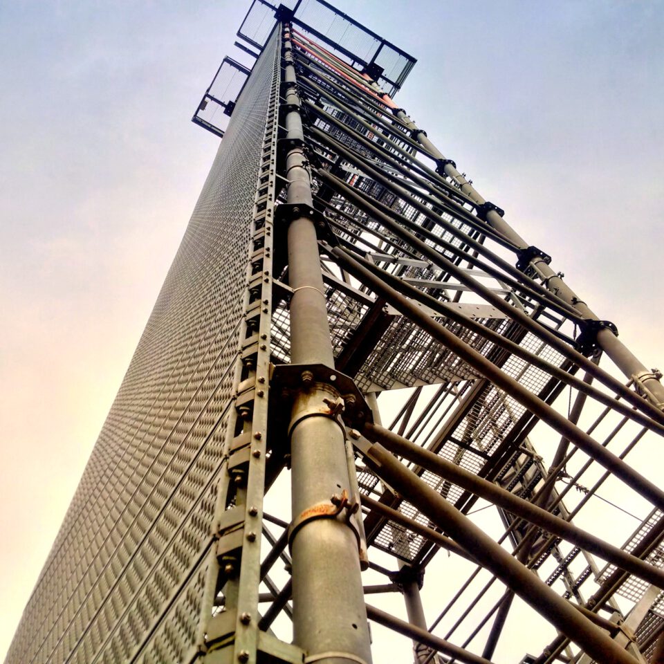 Das Bild zeigt eine Nahaufnahme aus der Froschperspektive auf einen hohen Metallkonstruktion, ein Kommunikationssturm. Die Struktur besteht aus zahlreichen Metallträgern und -streben, teilweise mit Gitterplattformen versehen. Im Hintergrund ist ein klarer Himmel mit einem Farbverlauf von hellem zu dunklerem Blau zu sehen.