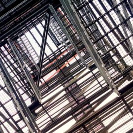 Das Bild zeigt eine Metallkonstruktion, ein Treppenaufgang aus der Froschperspektive. Mehrere Ebenen mit gitterartigen Plattformen erzeugen ein geometrisches Muster, das sich zur Bildmitte hin verjüngt.