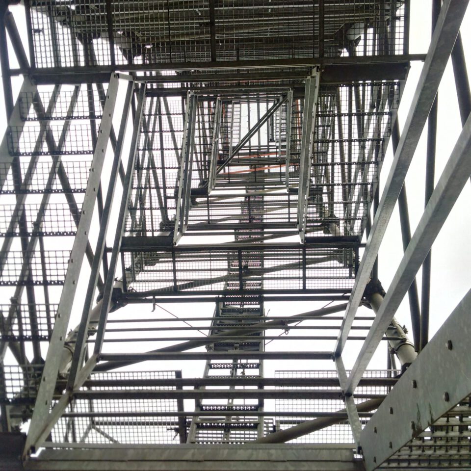 Das Bild zeigt die Unterseite einer Metallkonstruktion, vermutlich einen Turm aus der Froschperspektive. Die Perspektive erzeugt eine Reihe konzentrischer rechteckiger Rahmen aus Metallstreben und Gittern, die den Blick nach oben durch die Struktur lenken.