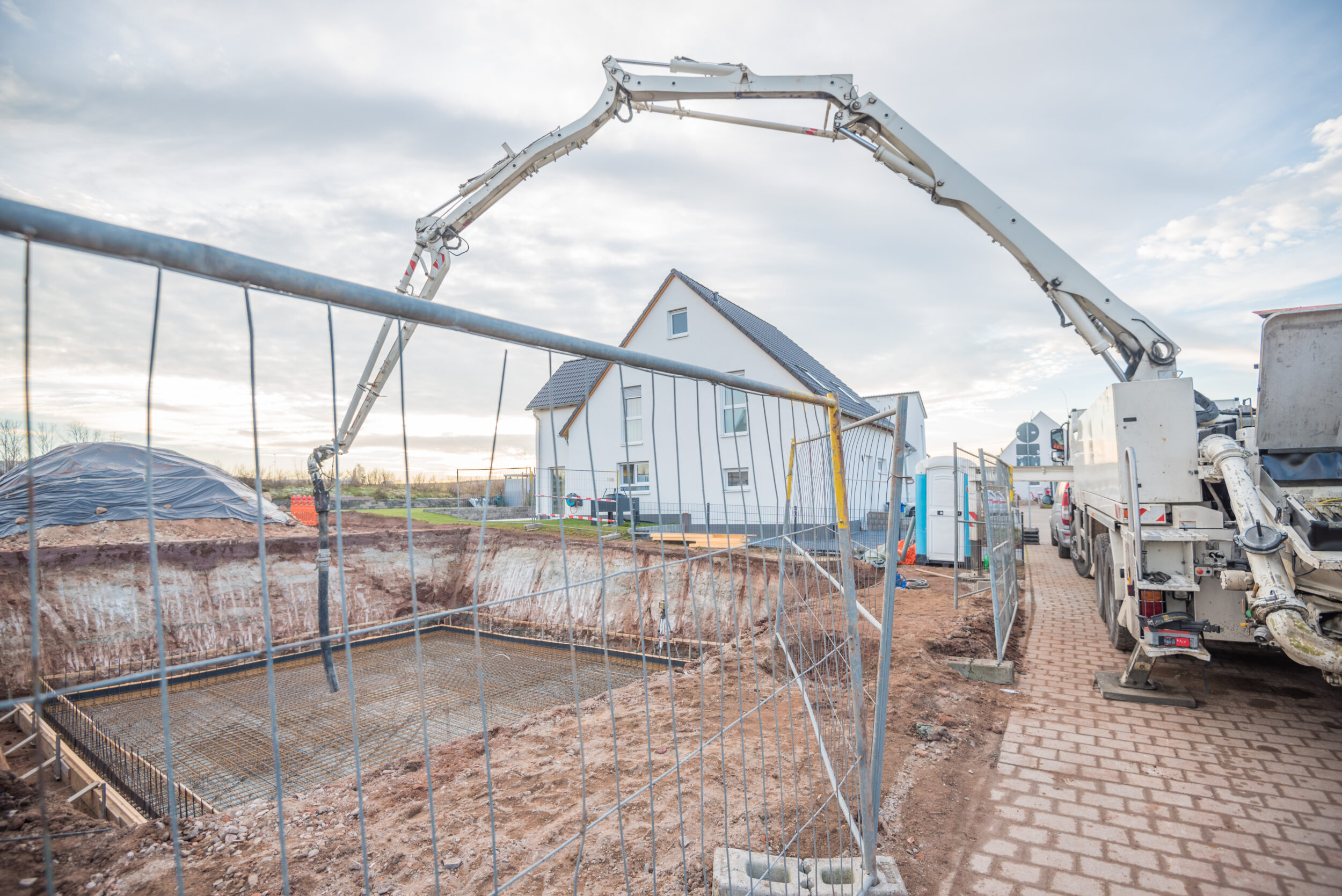 Das Bild zeigt eine Baustelle mit einem großen Betonpumpenfahrzeug, dessen Ausleger über ein abgesperrtes Fundamentbereich reicht. Im Hintergrund steht ein modernes weißes Haus mit dunklem Dach. Auf der Baustelle sind verschiedene Baumaterialien und Geräte verteilt, der Himmel ist teilweise bewölkt.