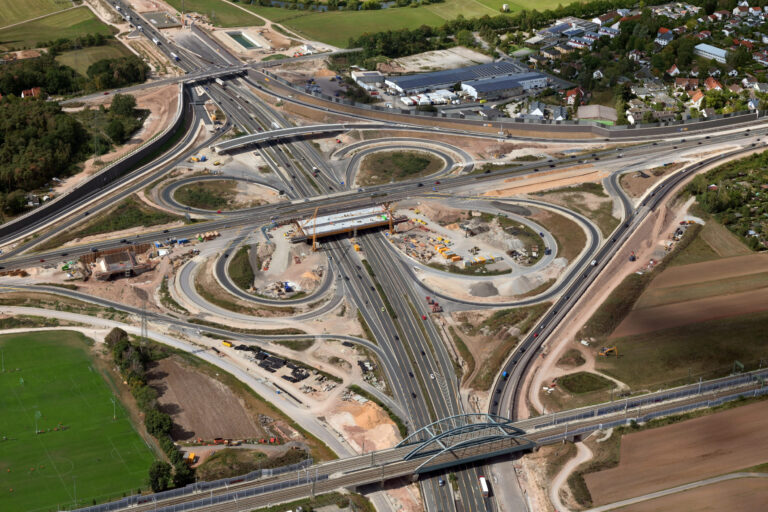 Das Bild zeigt eine Großbaustelle am Autobahnkreuz A3 mit mehreren Fahrspuren und Rampen. In der Mitte des Kreuzes sind Bauarbeiten im Gange, sichtbar durch Baumaschinen und Geräte. Umgeben ist das Areal von Feldern, Wohngebieten und Industrieanlagen.