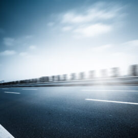 Das Bild zeigt einen Abschnitt einer Asphaltstraße mit deutlich sichtbaren Fahrbahnmarkierungen. Weiße gestrichelte Linien trennen die Fahrspuren, und am linken Rand verläuft eine durchgezogene weiße Linie. Im Hintergrund sind Bauwerke und ein teilweise bewölkter Himmel zu erkennen.