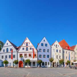 Malerischer Stadtplatz in Weiden mit einer Reihe traditioneller Gebäude mit Giebeldächern, einem markanten Kirchturm auf der linken Seite und mehreren Außensitzbereichen mit Tischen und Sonnenschirmen bei sonnigem Wetter.