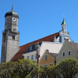 Malerische Stadtansicht in Weilheim mit einem markanten Uhrturm mit grüner Kuppel, umgeben von bunten Gebäuden mit roten Ziegeldächern, Bäumen im Vordergrund und blauem Himmel im Hintergrund.