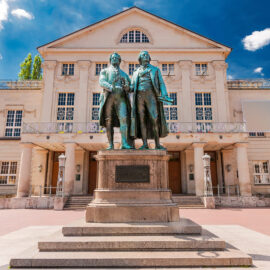 Ansicht des Goethe-Schiller-Denkmals in Weimar mit bronzenen Statuen der beiden Dichter auf einem Sockel vor dem Deutschen Nationaltheater mit klassizistischer Architektur; umgeben von Bäumen und weiteren Gebäuden bei leicht bewölktem Himmel.